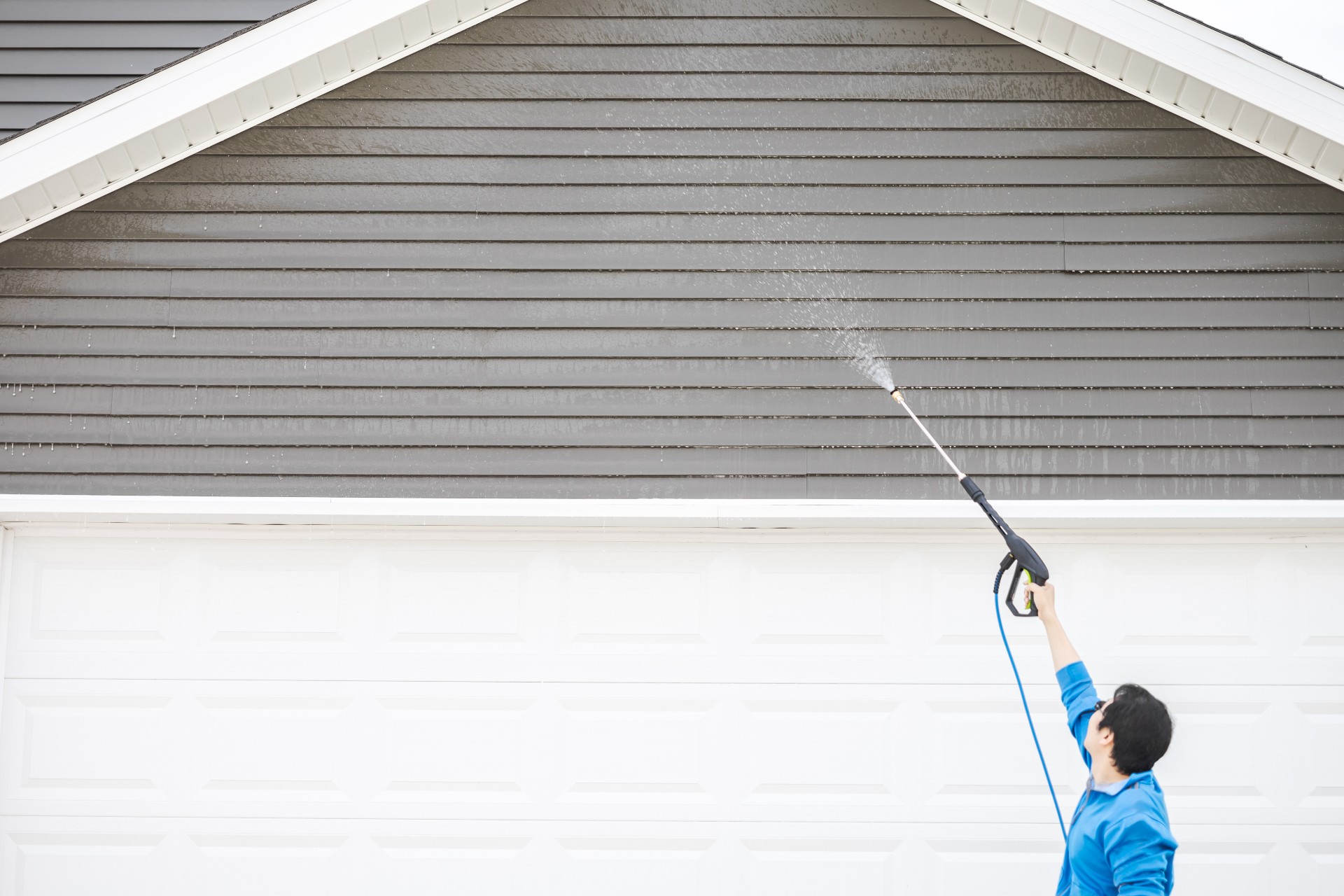 Man cleaning dirt and grime from exterior siding under the roof with a pressure washer.